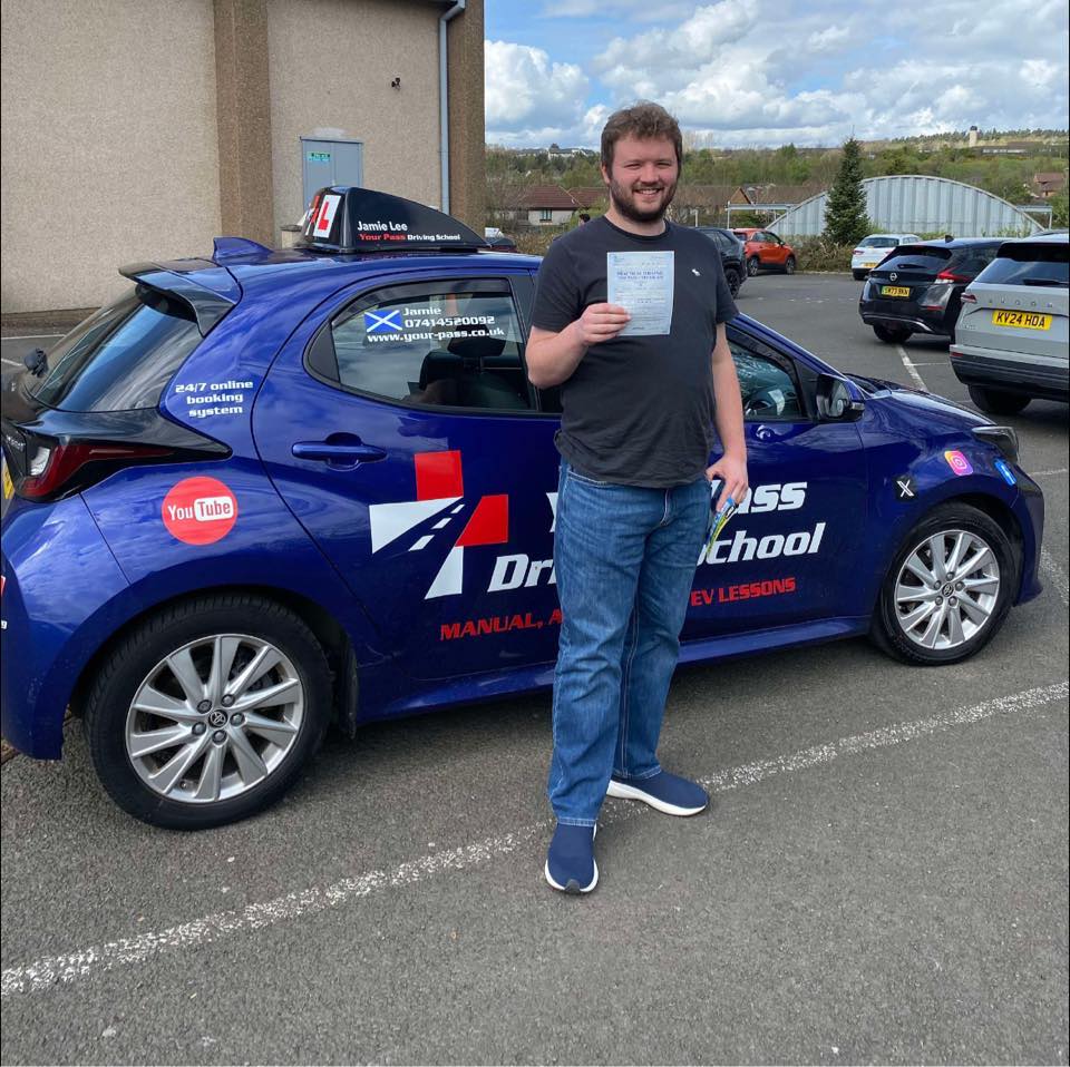 A smiling man stands proudly holding his driving test pass certificate next to a blue Your Pass Driving School car, which is branded with logos for manual, automatic, and EV lessons.