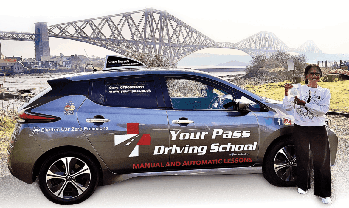 Smiling woman holding her driving test pass certificate stands next to a branded Your Pass Driving School electric car, with the Forth Bridge visible in the scenic background